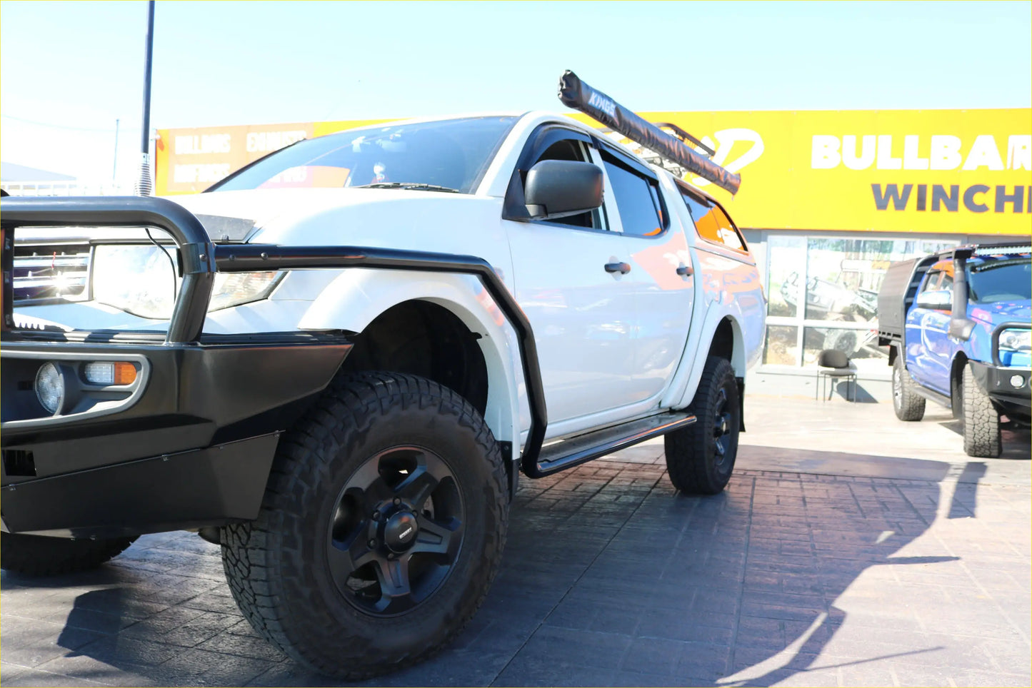 White mitsubishi triton with black rockarmor steel rockslider side steps and rugged off-road tires shown from the back