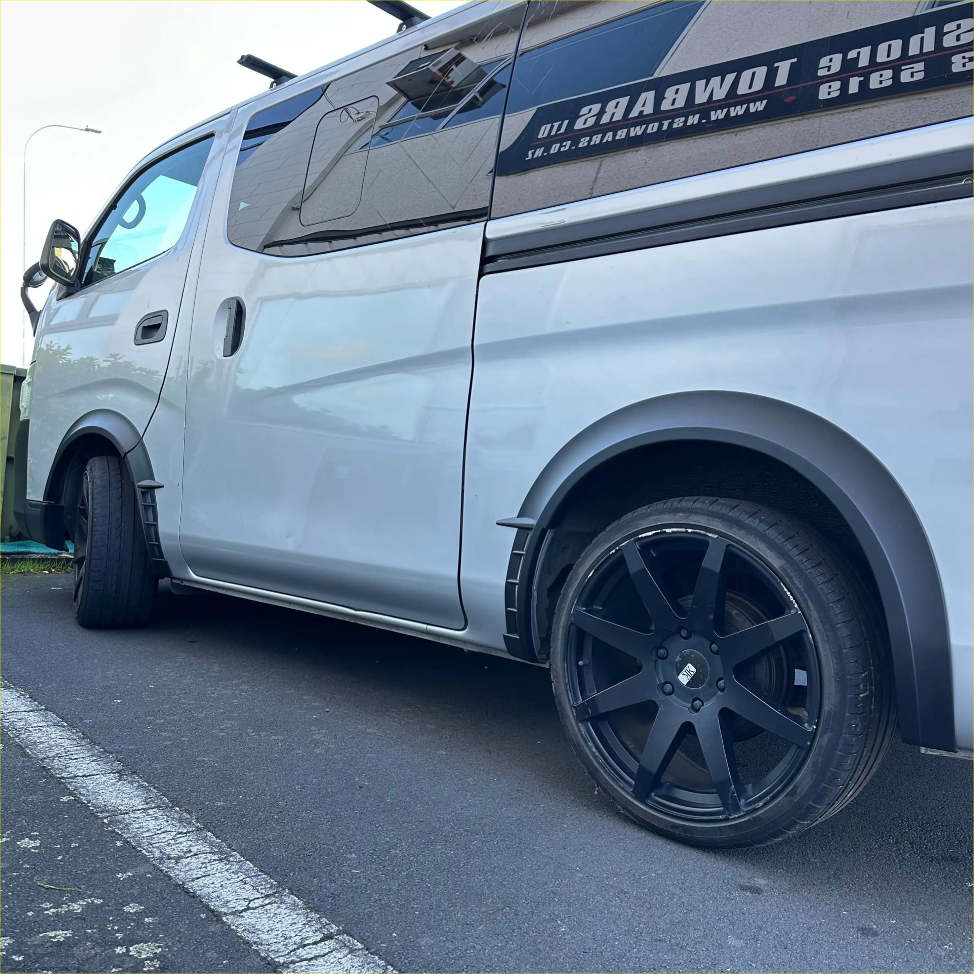 Silver nissan nv350 caravan with matte sport style fender flare and black alloy wheels shown from the back side